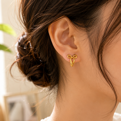 A close-up of a woman's ear wearing a small gold bow-shaped stud earring, with her hair in a low bun and a white top visible against a softly blurred indoor background.