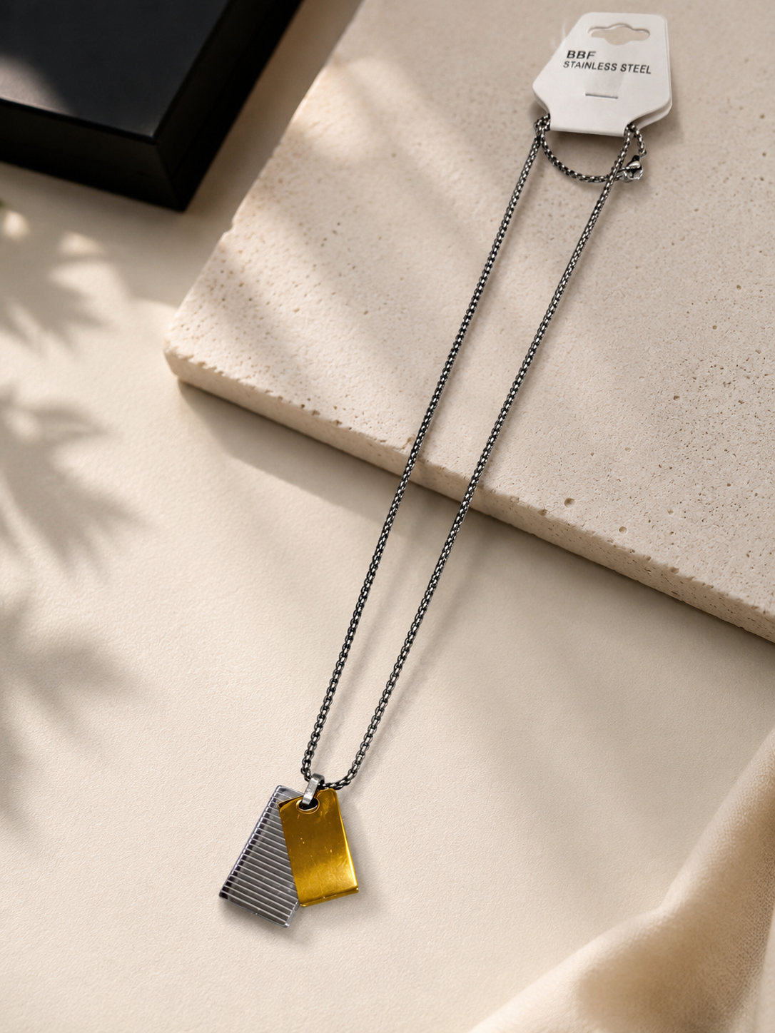 A silver-toned chain necklace with two geometric dog tag pendants, one textured silver and one brushed gold, displayed on a beige stone