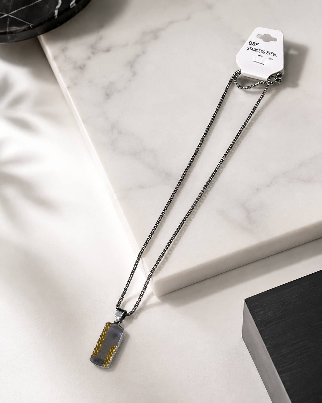 A stainless steel dog tag necklace with a textured gold and silver patterned pendant, displayed on a white marble block with its retail tag attached.