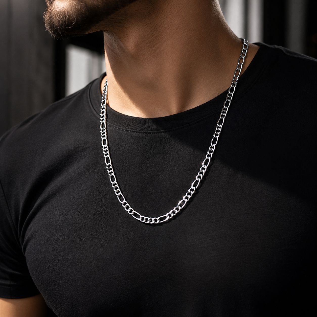A close-up of a man wearing a silver Figaro chain necklace over a black t-shirt.