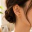 A close-up of a woman's ear wearing a small gold bow-shaped stud earring, with her hair in a low bun and a white top visible against a softly blurred indoor background.