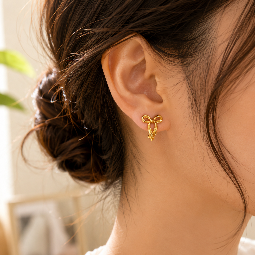 A close-up of a woman's ear wearing a small gold bow-shaped stud earring, with her hair in a low bun and a white top visible against a softly blurred indoor background.